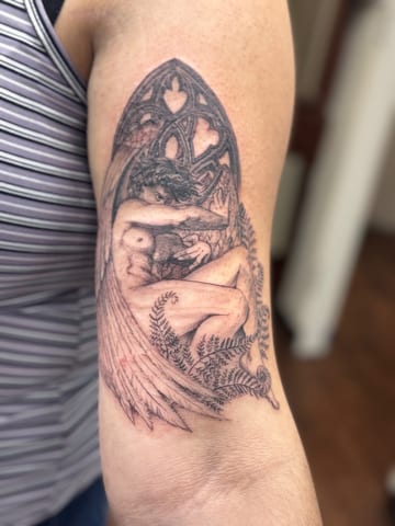 A delicate black and grey tattoo of an angel with wings and ferns on the upper arm, featuring intricate illustrative details — by Sandra Price, los angeles