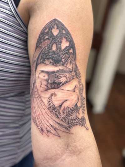 A delicate black and grey tattoo of an angel with wings and ferns on the upper arm, featuring intricate illustrative details — by Sandra Price, los angeles