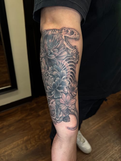 A black and grey dinosaur skeleton intertwined with flowers on the forearm, featuring detailed illustrative style — by Sandra Price, los angeles
