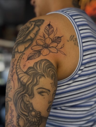 A black and grey tattoo of a woman with horns and flowers on the upper arm in an illustrative style — by Maddie Ramirez, los angeles