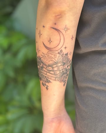 A delicate black and grey tattoo of a stack of books with a crescent moon and stars on the forearm, featuring fine line floral details — by Sandra Price, los angeles