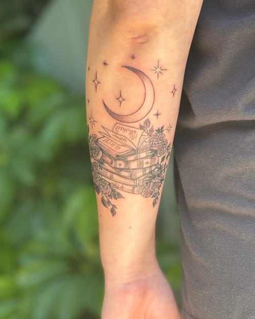 A delicate black and grey tattoo of a stack of books with a crescent moon and stars on the forearm, featuring fine line floral details — by Sandra Price, los angeles
