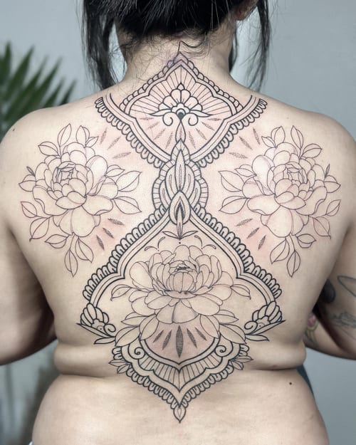 An ornate black and grey floral design tattoo covering the back, featuring intricate ornamental patterns and delicate flower outlines — by Melanie Benoit, los angeles