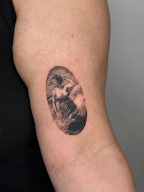 black and grey oval vignette of a woman in ocean waves on the inner upper arm — by lunayflorestattoo, los angeles