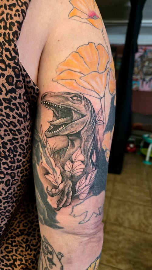 A Neo-Traditional tattoo featuring a dinosaur with open jaws surrounded by flowers on the upper arm, combining blackwork with subtle color accents — by Dretty, los angeles