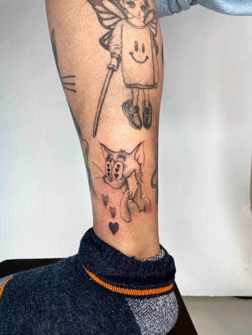 A black and grey cartoon-style tattoo of a cat character on the leg, featuring playful expressions and small heart accents — by Brian Kies, los angeles