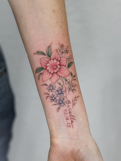 A delicate color floral tattoo on the forearm featuring a mix of flowers with fine line details and script lettering — by Melanie Benoit, los angeles