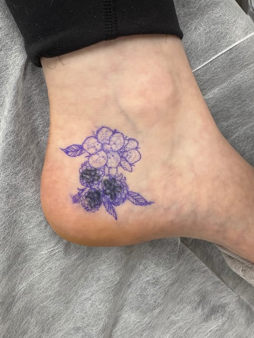 A delicate black and grey fine line tattoo of a flower and berry cluster on the ankle — by Sandra Price, los angeles