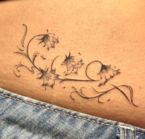 A delicate fine line floral tattoo on the hip, featuring black and grey ink with intricate flower designs — by NJ TATTOOZ, los angeles