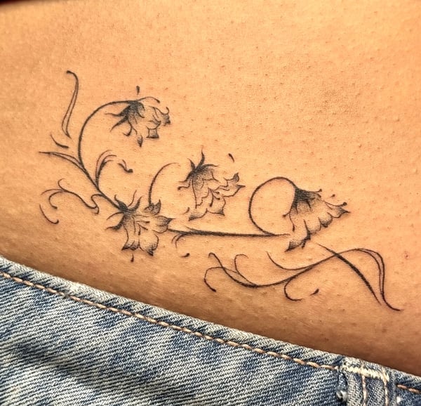 A delicate fine line floral tattoo on the hip, featuring black and grey ink with intricate flower designs — by NJ TATTOOZ, corona, ca
