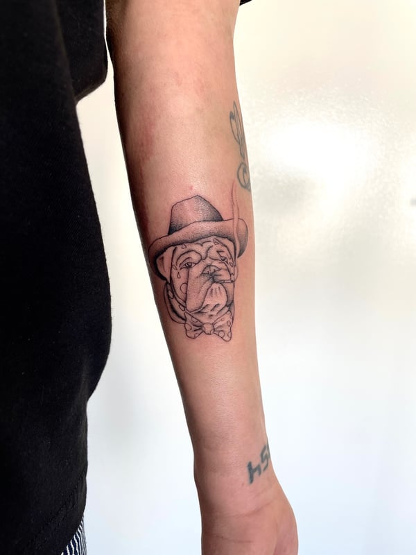 A whimsical black and grey bulldog wearing a hat tattooed on the forearm with clean linework — by Brian Kies, los angeles