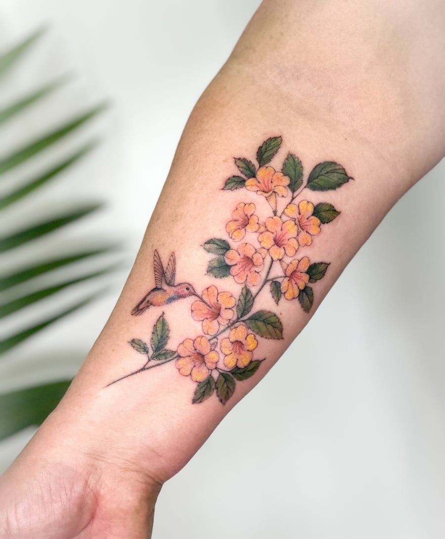 A delicate color tattoo of a hummingbird and flowers on the forearm, featuring fine line botanical details — by Melanie Benoit, los angeles