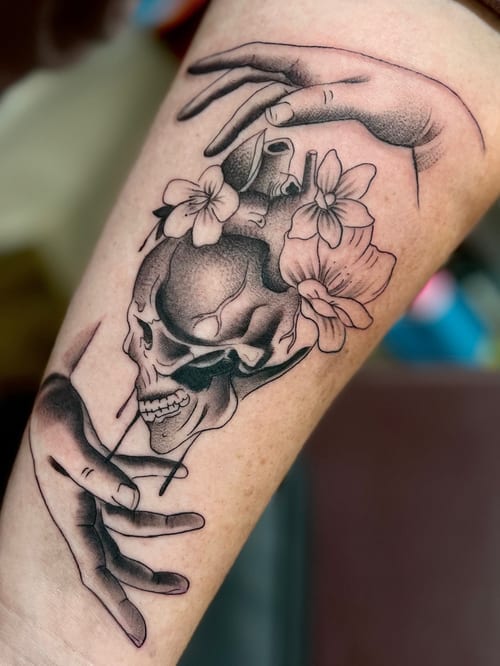 A realistic black and grey tattoo on the thigh featuring a skull intertwined with flowers and hands, showcasing detailed shading and depth — by Josue Bravo, los angeles