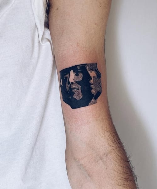 An abstract blackwork tattoo on the upper arm featuring stylized faces within a dark, irregular shape — by Level tattoo, los angeles