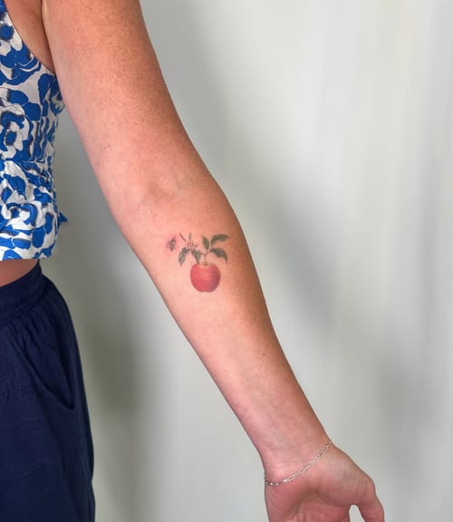A delicate color tattoo of an apple with leaves on the forearm, done in an illustrative style — by Melanie Benoit, los angeles