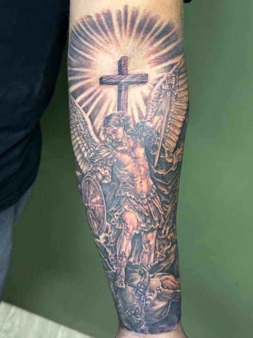 A detailed black and grey realism tattoo of an angel with a cross on the forearm, featuring intricate shading and religious symbolism — by Humilde tattoos, los angeles