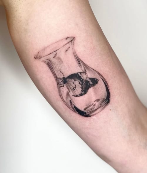 A black and grey realism tattoo of a butterfly inside a glass vase on the forearm, featuring detailed shading and a reflective effect — by Level tattoo, los angeles