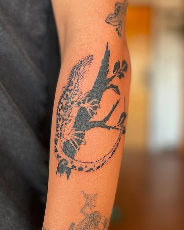 A black and grey illustrative tattoo of a lizard on a branch located on the arm — by Dylan Aiello, los angeles
