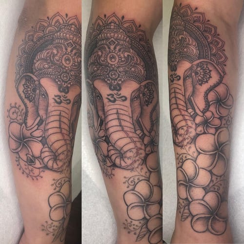 A detailed black and grey tattoo of Ganesha with an ornamental mandala design on the forearm, featuring floral elements — by Sandra Price, los angeles