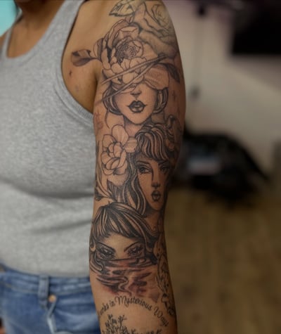 A black and grey tattoo of three women's faces with floral elements on the arm, in an illustrative blackwork style — by Maddie Ramirez, los angeles