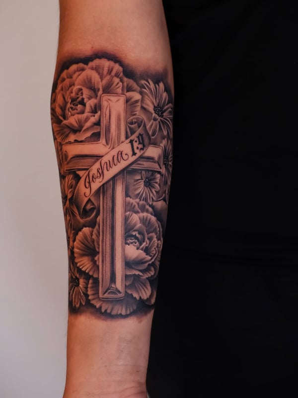 A black and grey tattoo of a cross with flowers and script on the forearm, featuring realistic shading and intricate details — by Johnny Ayala, fontana, ca