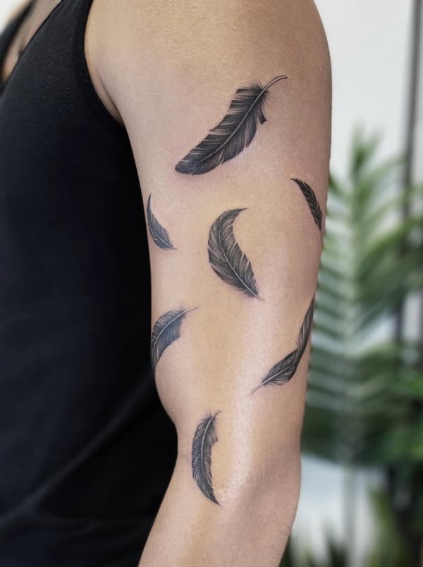 A series of black and grey fine line feathers on the upper arm, arranged in a flowing pattern with delicate shading — by Jian Liang, los angeles