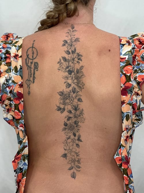 A delicate black and grey floral tattoo running vertically along the spine, featuring detailed flowers and leaves — by Melanie Benoit, los angeles