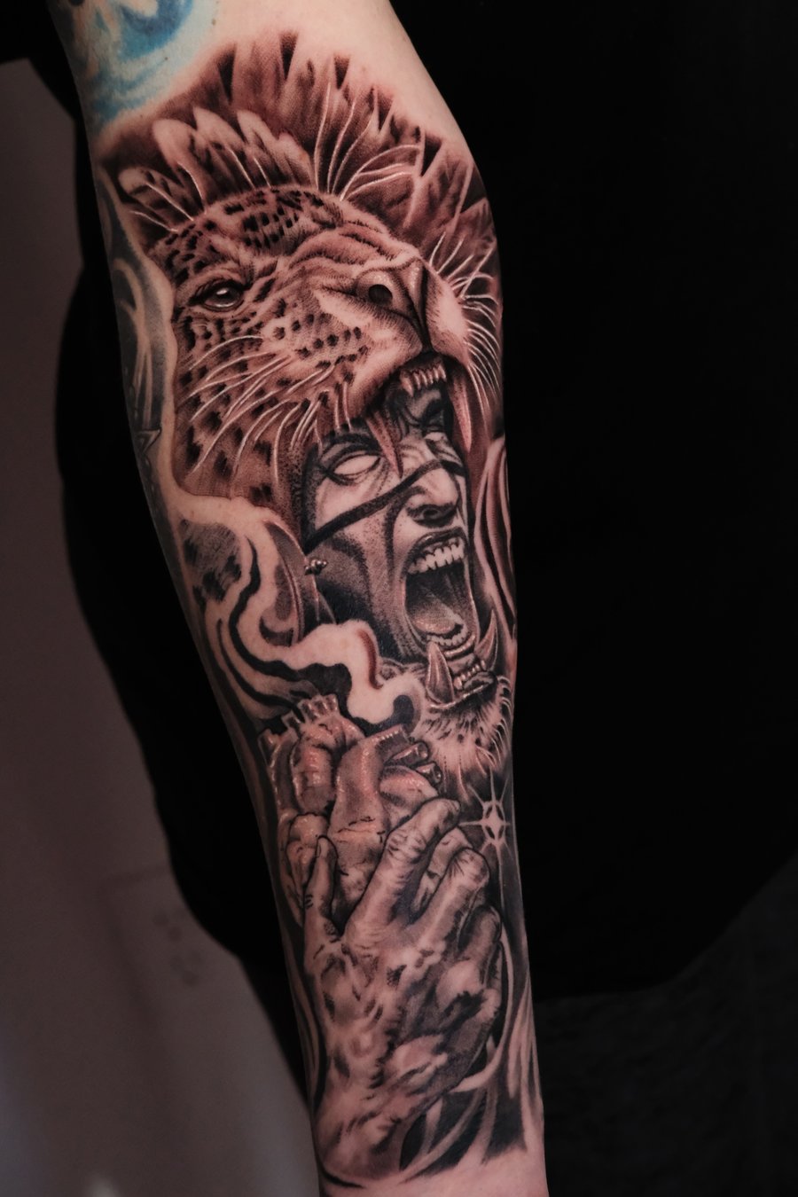 A detailed black and grey realism tattoo on the forearm depicting a roaring lion above a screaming face with a hand holding a heart — by Johnny Ayala, los angeles