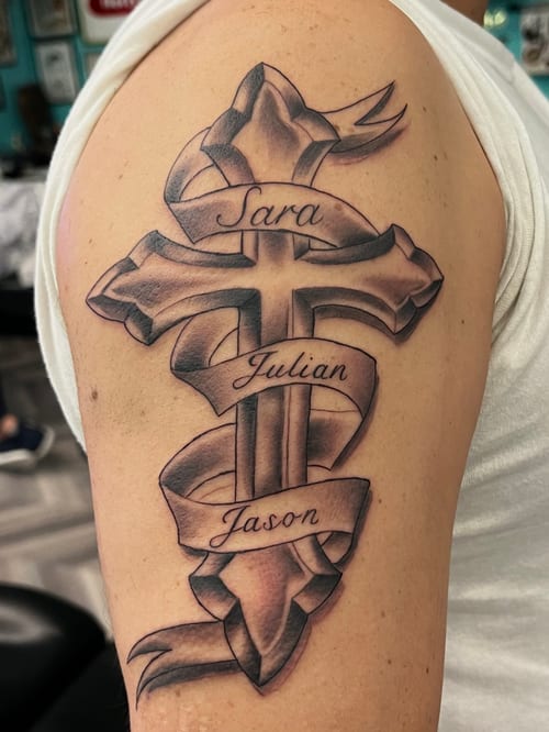 A black and grey cross tattoo on the upper arm, featuring a ribbon with names in script lettering — by Josue Bravo, los angeles