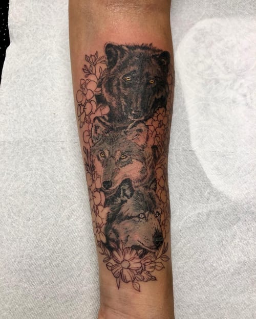 A black and grey tattoo featuring a wolf and bear surrounded by flowers on the forearm, showcasing realistic detail and bold shading — by Sandra Price, los angeles