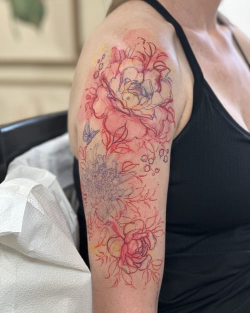 A delicate watercolor floral tattoo on the upper arm featuring colorful flowers and a butterfly — by Melanie Benoit, los angeles
