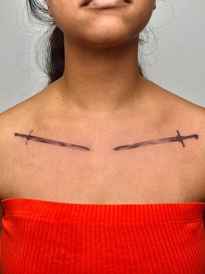 A minimalist black ink tattoo of crossed swords on the collarbone, featuring simple linework — by Dylan Aiello, los angeles