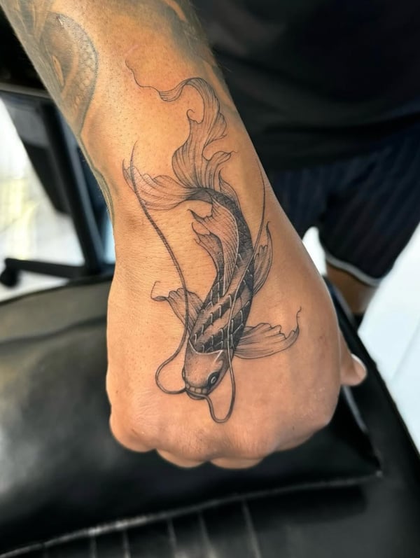 A fine line black and grey tattoo of a koi fish on the hand, featuring delicate linework and flowing fins — by otaybeatdown, orange, ca