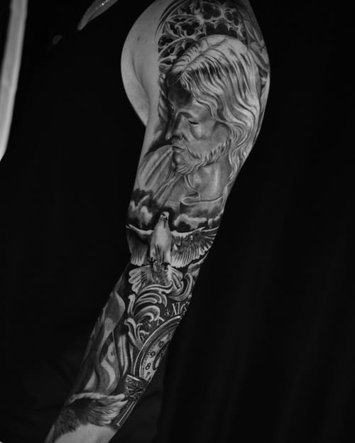 A black and grey full sleeve tattoo featuring a realistic portrait of Jesus Christ, a dove, and a clock, showcasing intricate details and spiritual themes — by Johnny Ayala, los angeles