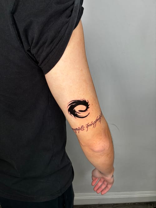 A black and grey tattoo on the outer arm featuring a bold blackwork design with script lettering underneath — by Edgar Armas Rodriguez, los angeles