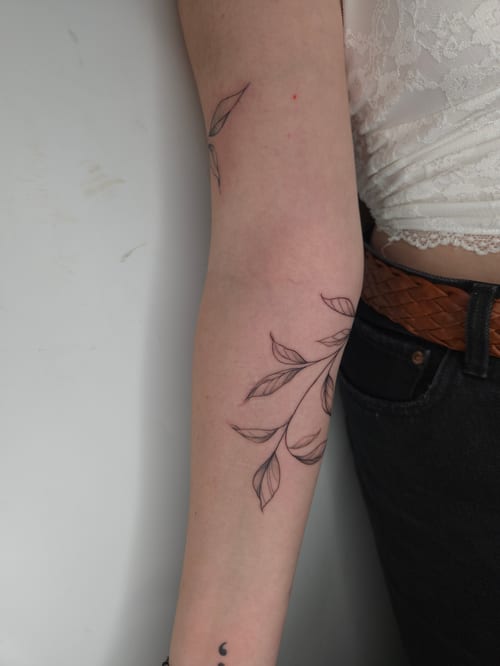 A minimalist fine line black and grey floral tattoo on the forearm, featuring delicate leaves and branches — by ashynucklestattoo, los angeles