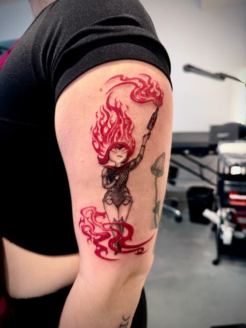 A colorful illustrative tattoo of a woman with flaming hair holding a paintbrush, located on the upper arm. The design features vibrant red and black tones with a dynamic, fiery effect — by zitronx…