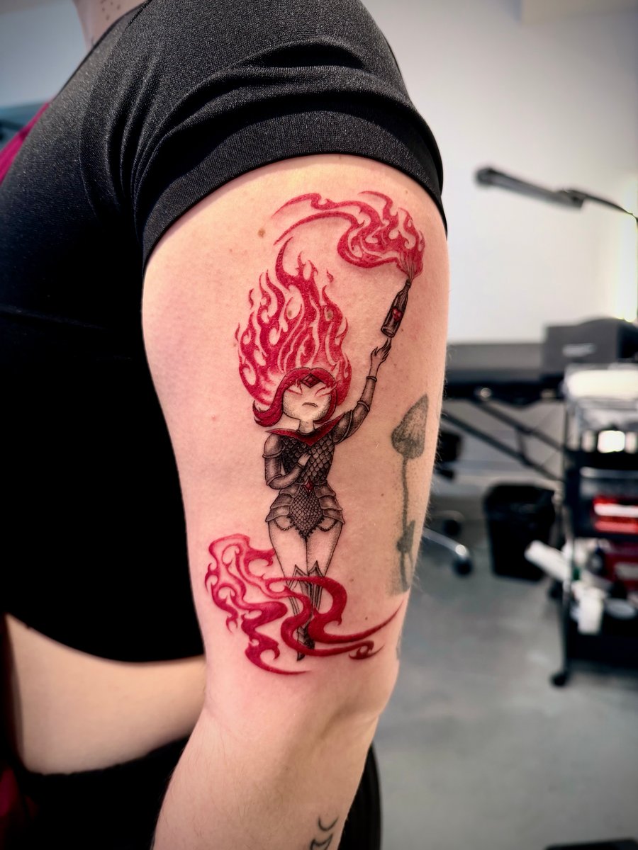 A colorful illustrative tattoo of a woman with flaming hair holding a paintbrush, located on the upper arm. The design features vibrant red and black tones with a dynamic, fiery effect — by zitronx…