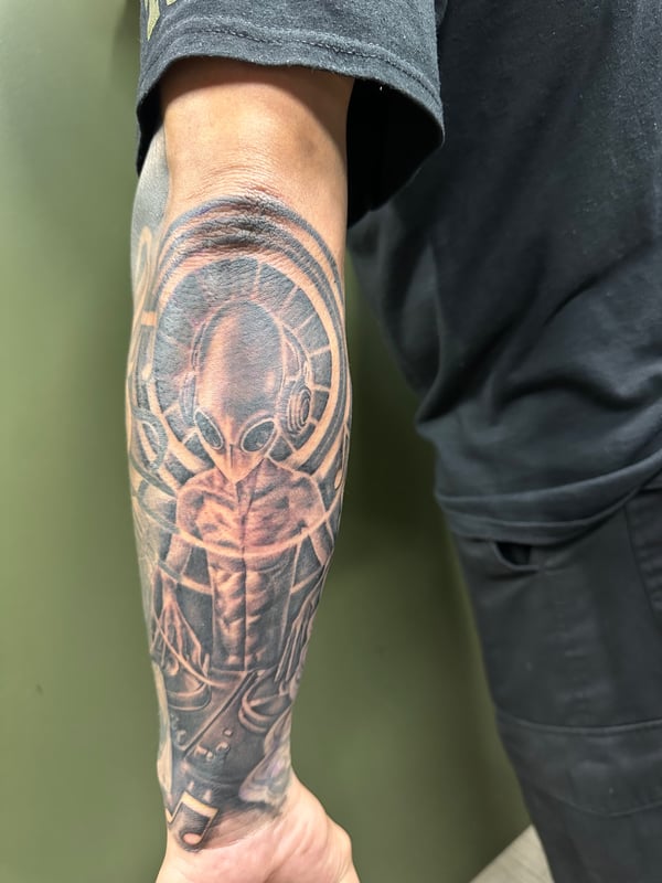 A black and grey realism tattoo of an alien DJ on the forearm, featuring detailed shading and a cosmic theme — by Humilde tattoos, los angeles