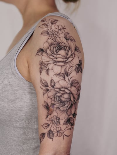 A delicate black and grey floral arrangement with a hummingbird on the upper arm, featuring fine line details and subtle shading — by Melanie Benoit, los angeles