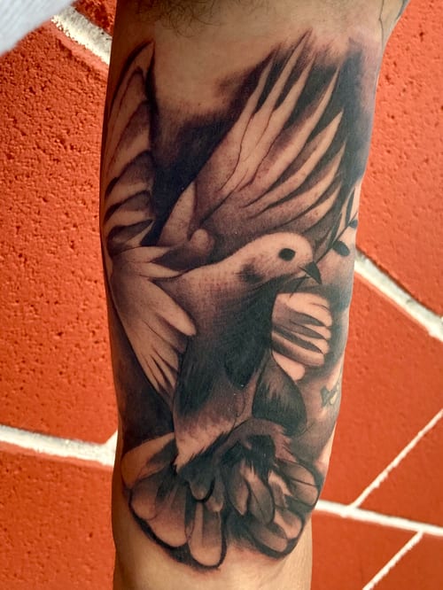 A realistic black and grey tattoo of a bird in flight surrounded by flowers on the forearm, showcasing detailed shading and depth — by Josue Bravo, los angeles