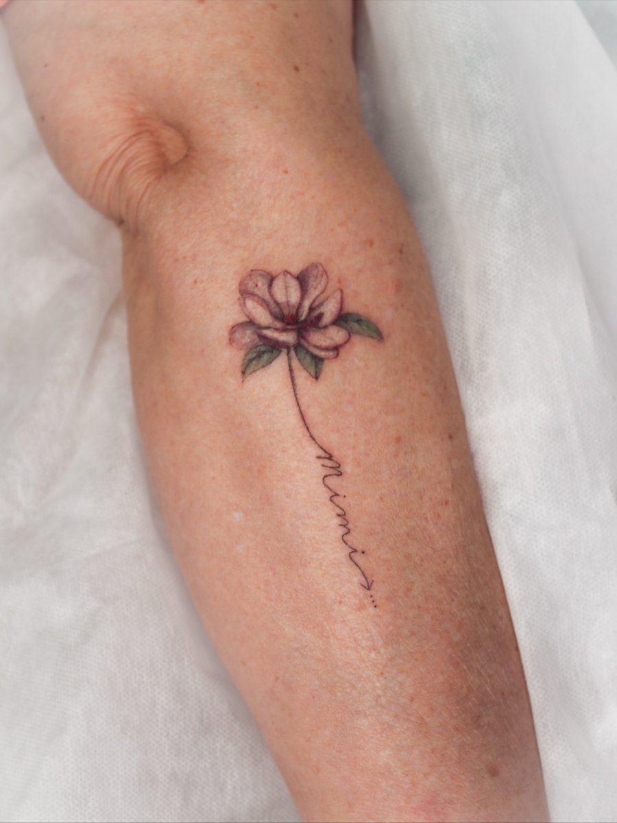 A delicate fine line flower tattoo with script on the forearm, featuring a minimalist design in color — by Melanie Benoit, los angeles