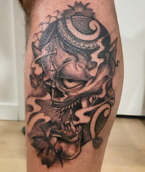 A black and grey dark art tattoo of a demonic skull with intricate patterns and shading on the leg — by Dretty, los angeles