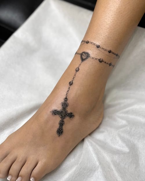 An ornamental black and grey tattoo of a cross with a beaded anklet design around the ankle, featuring intricate patterns and fine linework — by otaybeatdown, los angeles