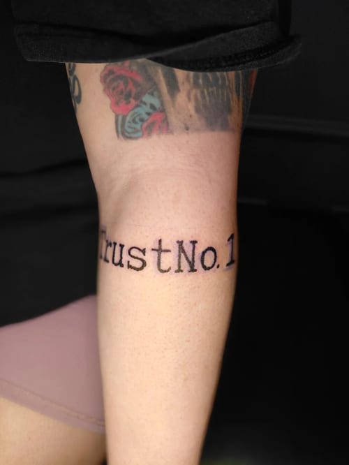 A black and grey lettering tattoo on the forearm with the text 'Trust No. 1' — by anoetic.designs.ultd, los angeles