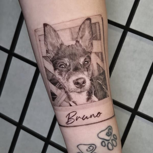 black and grey realism pet portrait of a dog inside a polaroid frame with paw prints on the forearm by Miguel Angelo, a tattoo artist in temecula, california