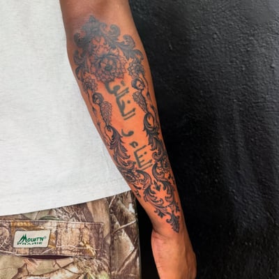 An ornate blackwork tattoo on the forearm featuring decorative script and floral elements — by Kiera Garvin, los angeles
