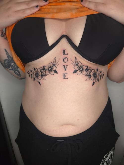 A black and grey tattoo on the stomach featuring the word 'LOVE' surrounded by floral designs on either side — by ashynucklestattoo, los angeles
