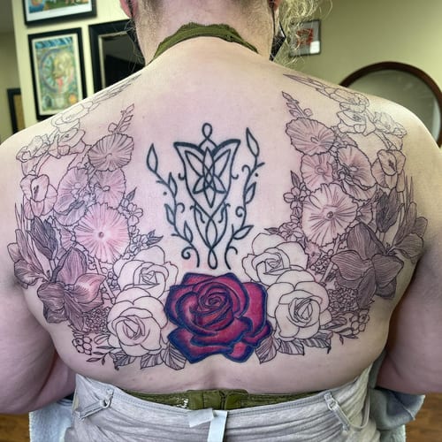 A large floral back tattoo featuring black and grey flowers with a central colorful rose and ornate linework — by Sandra Price, los angeles