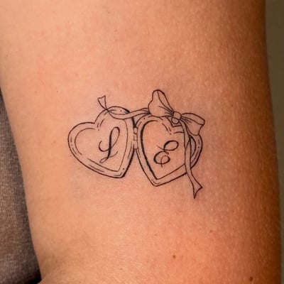 A delicate black and grey tattoo of two heart lockets with initials on the arm, featuring minimalist linework — by NJ TATTOOZ, los angeles